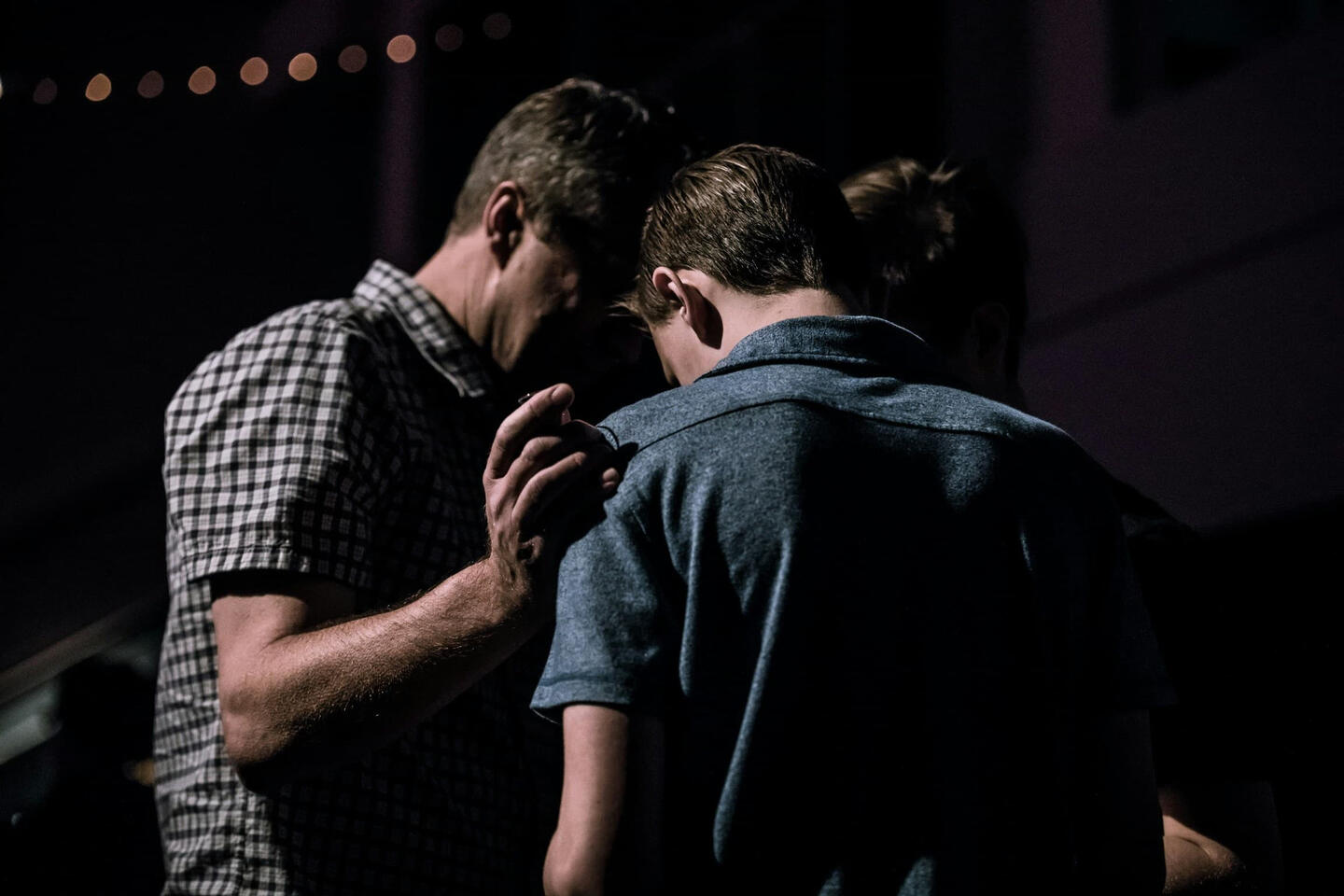 Two men in prayer together - one wearing a plaid shirt, placing a supportive hand on the shoulder of the man during an intimate moment of connection and spiritual support.